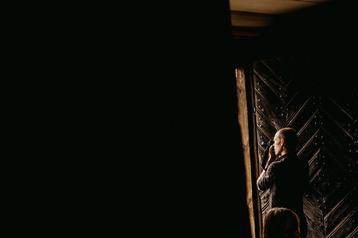 Person standing in shadows beside ornate wooden door in dramatic low light.