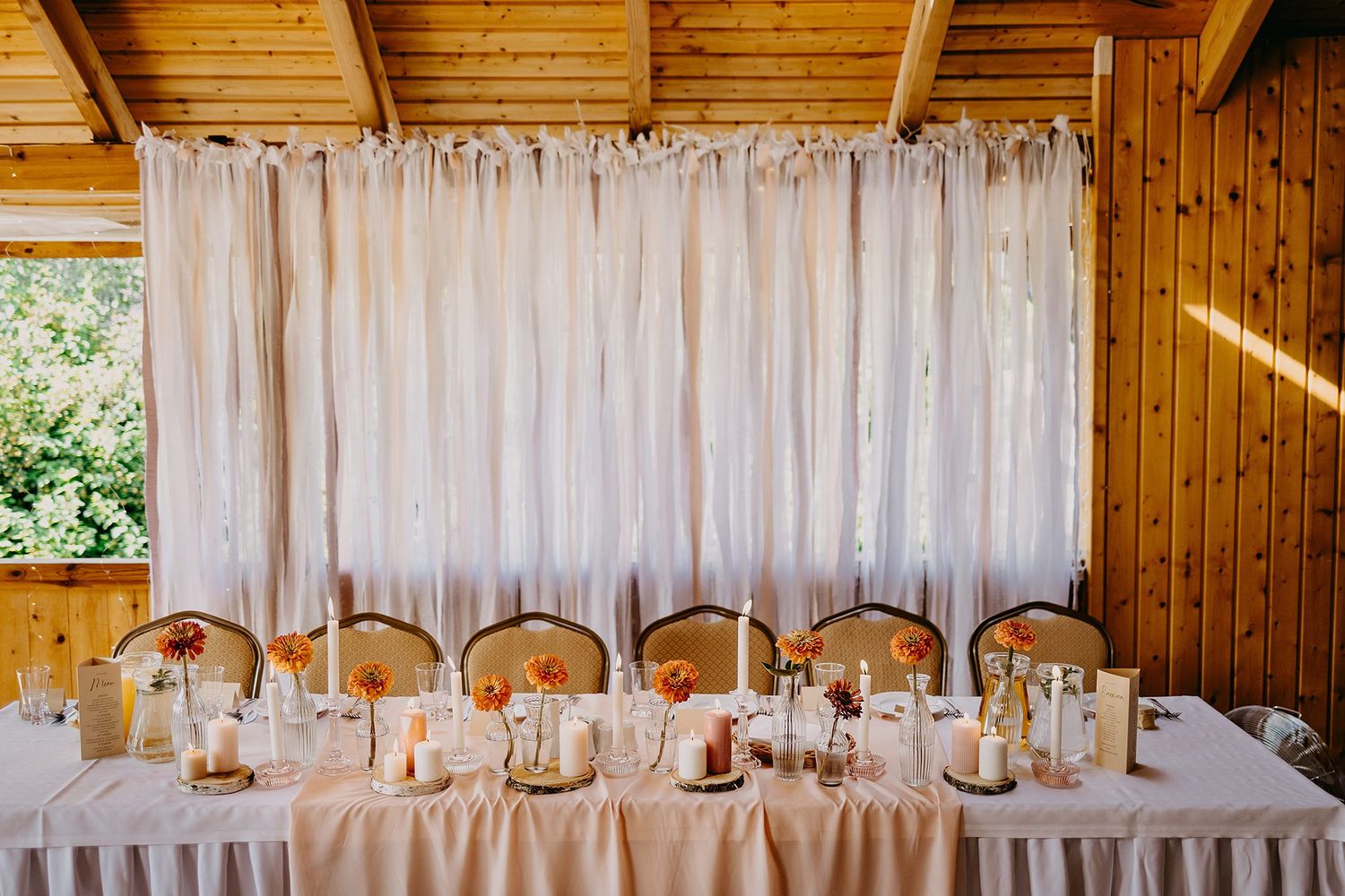 An elegant wedding head table set with candles, floral centerpieces, and a flowing white fabric backdrop in a wooden venue.