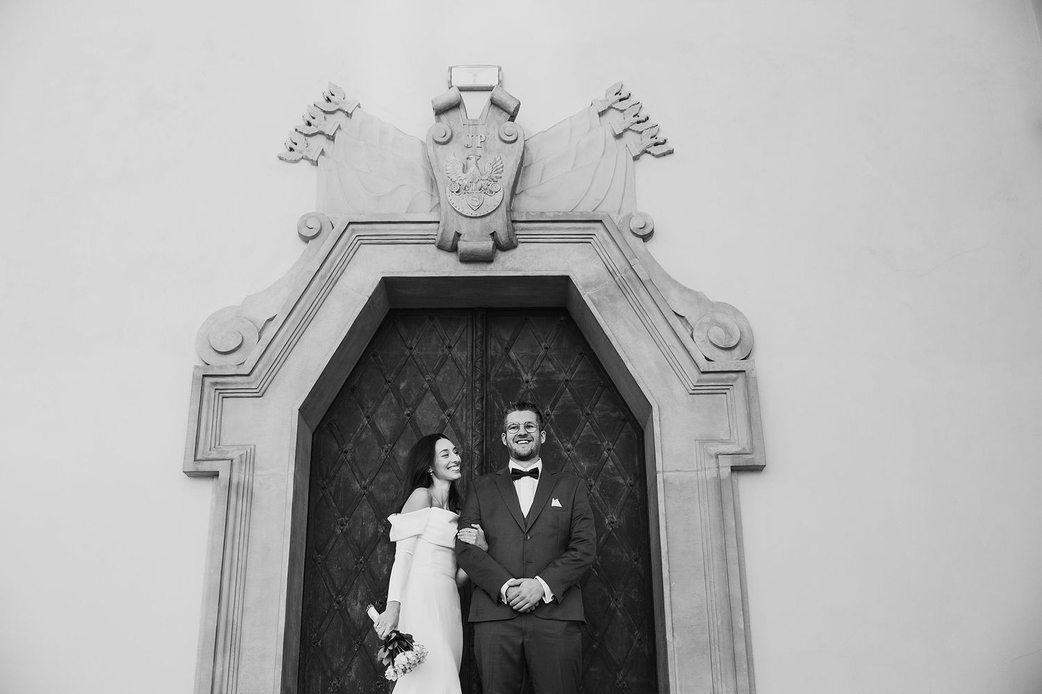 Black and white photo of bride and groom standing before ornate church door.
