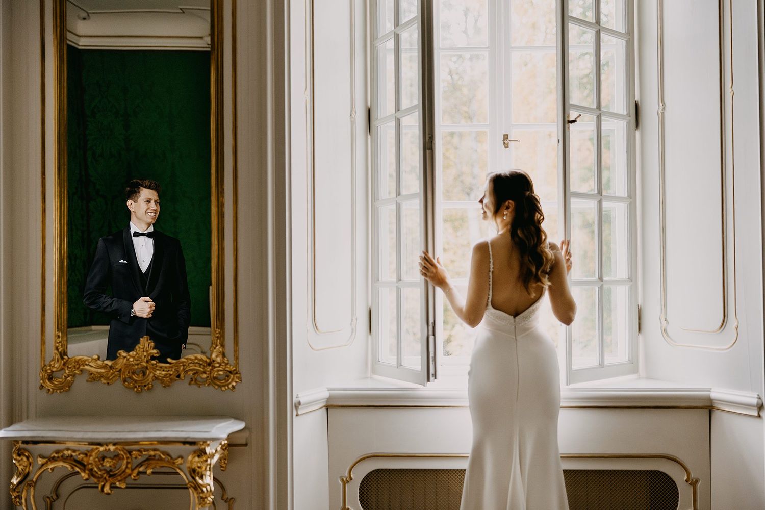 Bride in backless gown at window reflected in ornate gold mirror beside groom in tuxedo.