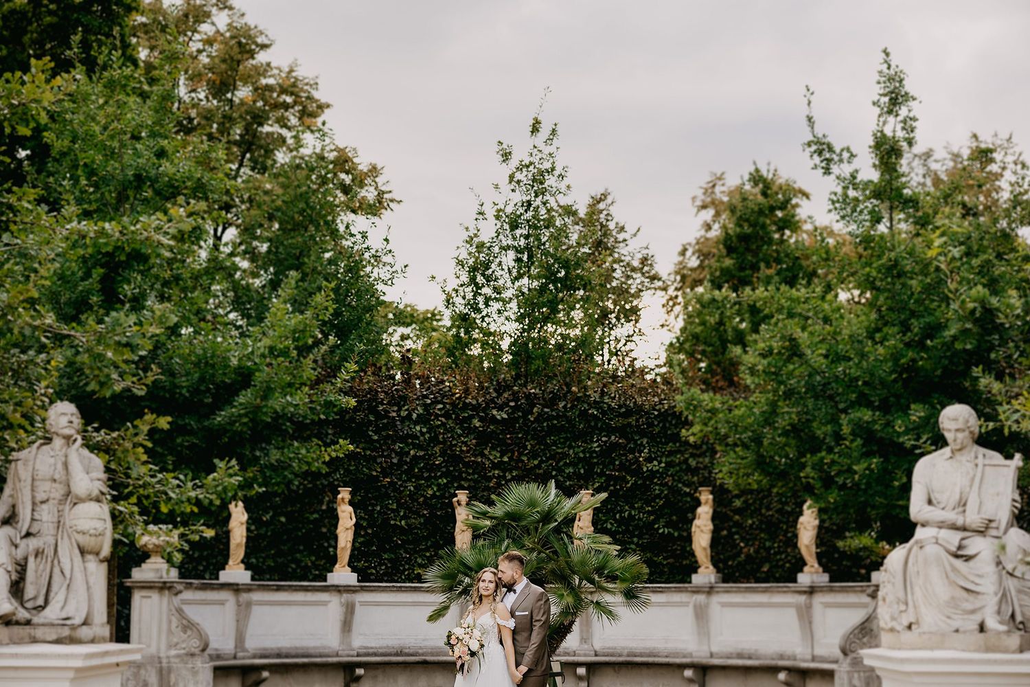 Elegant wedding couple posing between classical stone statues in formal garden with lush greenery.