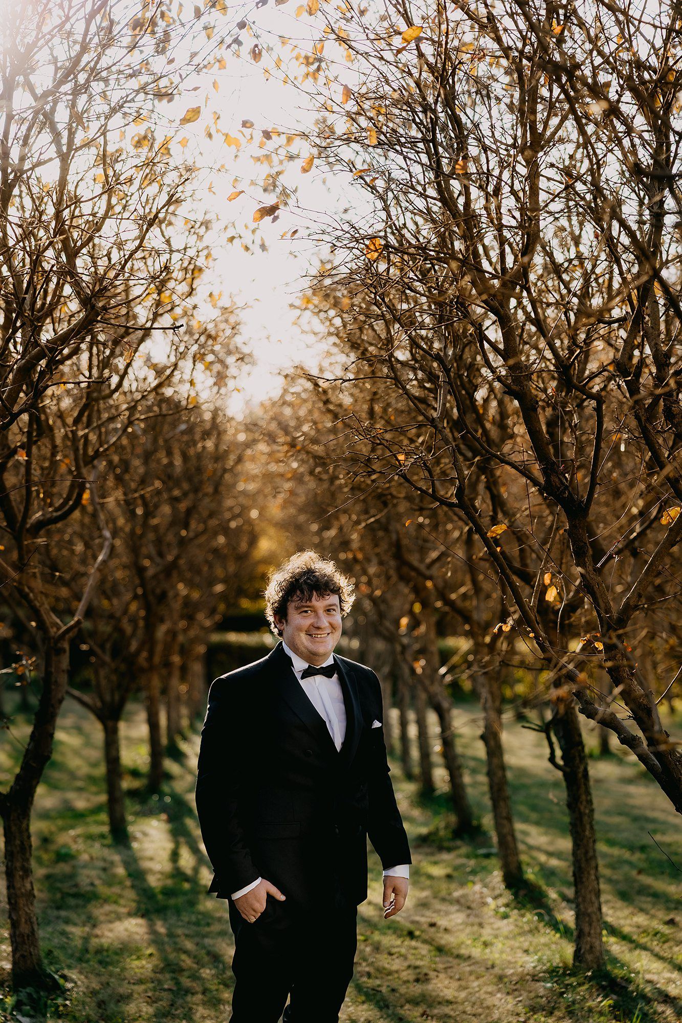Smiling groom in black tuxedo stands confidently among autumn bare trees in warm golden light.