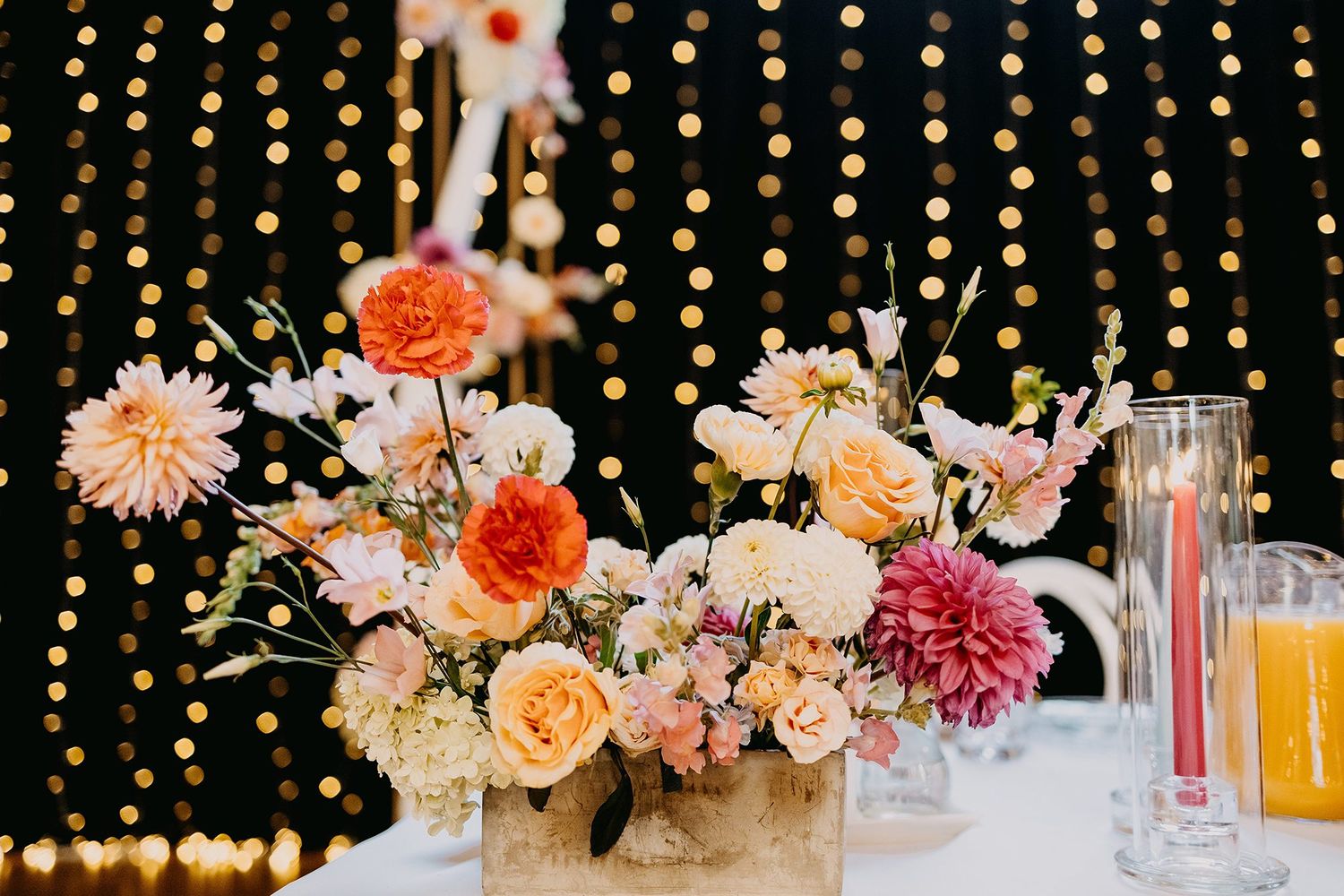 Colorful wedding centerpiece with peach roses, dahlias, and pink flowers in a wooden box against a bokeh light backdrop.