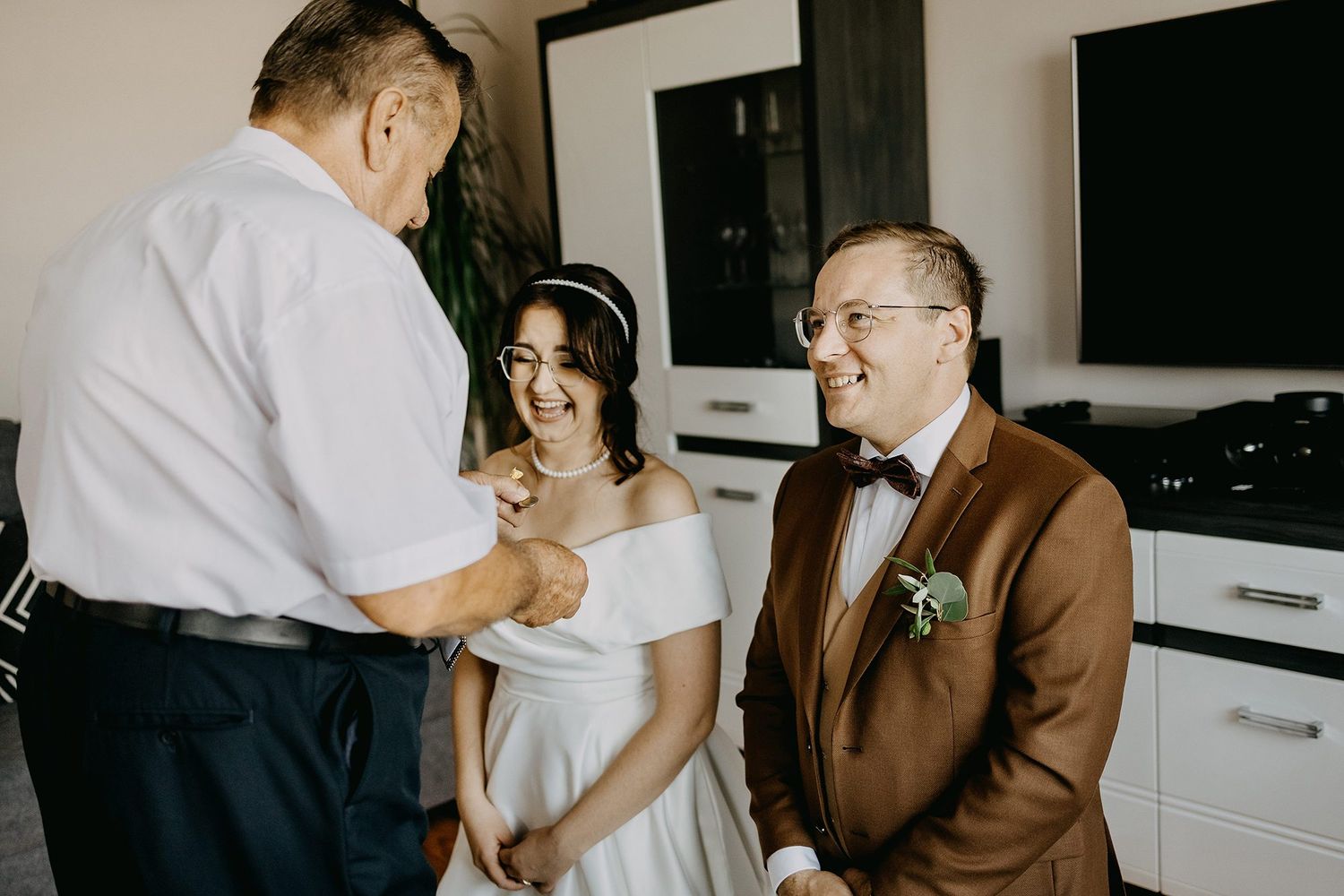Bride and groom in brown suit sharing a joyful moment with guest indoors before wedding.