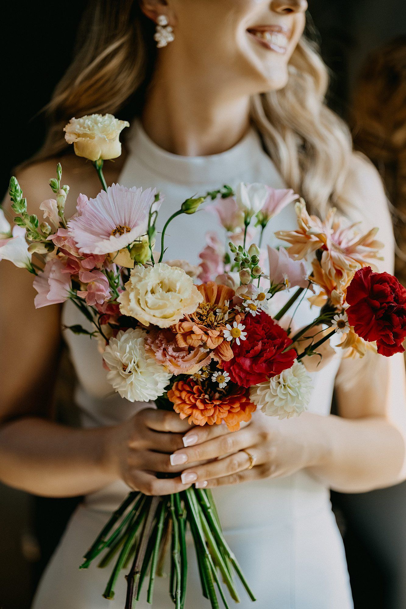 Smiling bride in white dress holding a vibrant wedding bouquet of pink, red, and orange flowers.