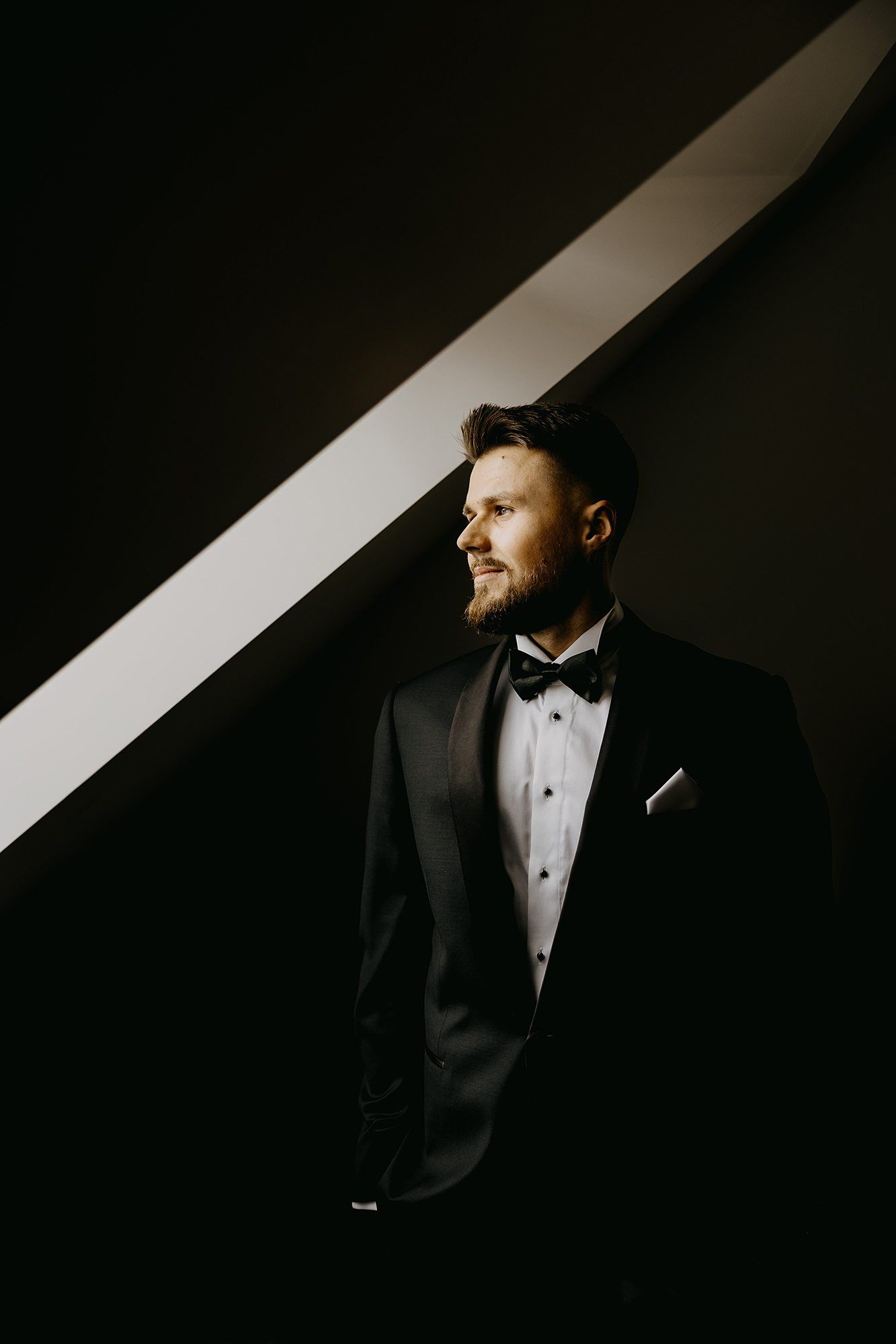 Groom in black tuxedo with bow tie standing in dramatic light and shadow.