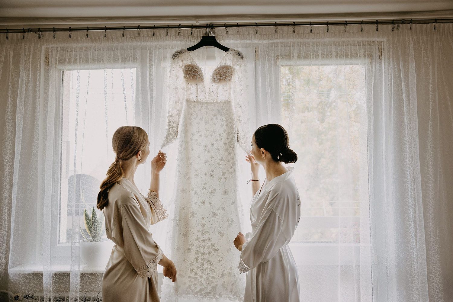 Two bridesmaids in robes admiring a beaded wedding gown hanging by a bright window.