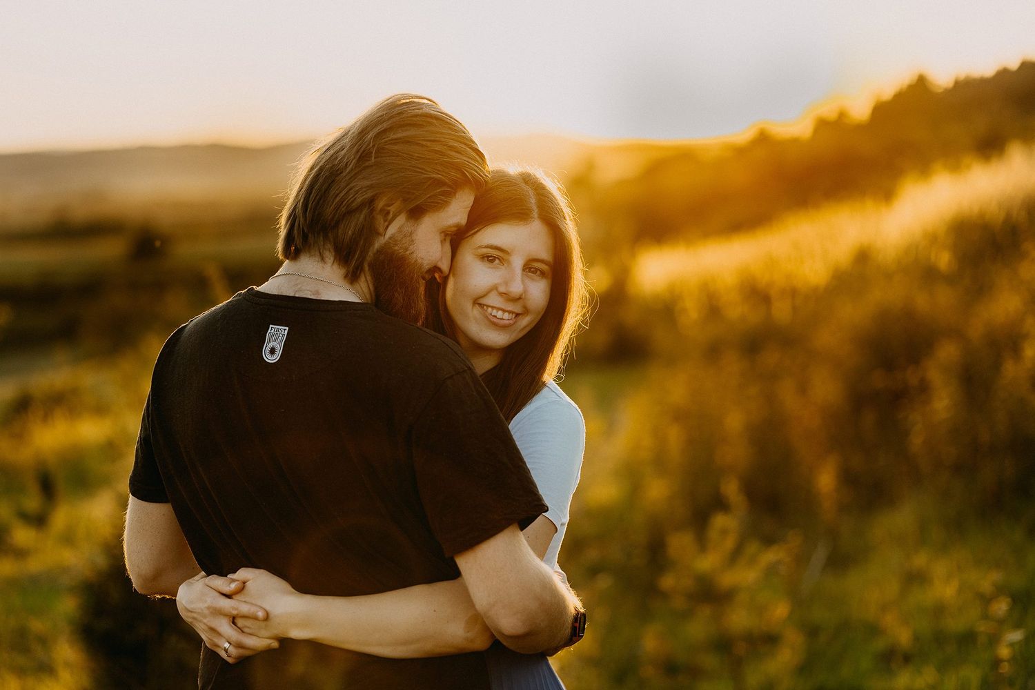 Couple embracing and smiling in a golden sunlit field at sunset, woman looking joyfully at camera.