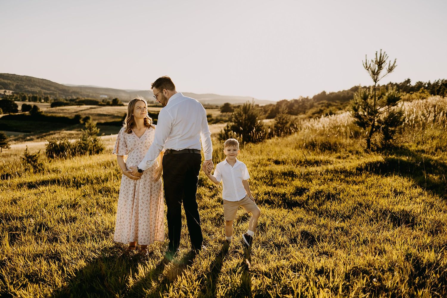 Pregnant woman in floral dress walking with partner and young son in sunny green meadow.
