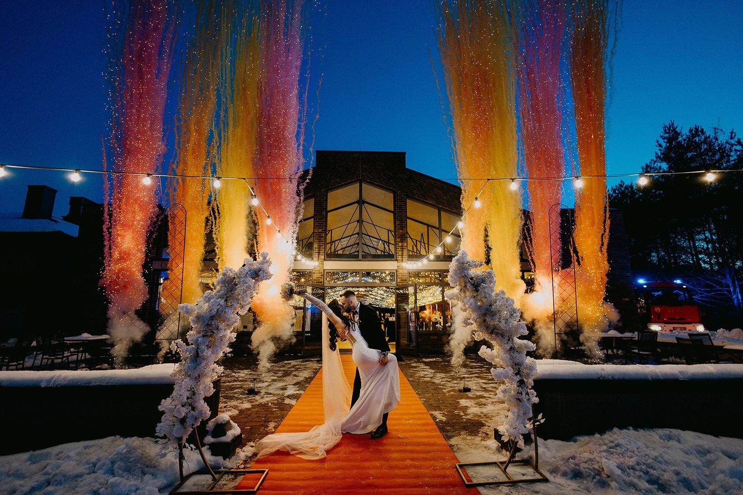 A bride and groom stand under vibrant colorful streamers on a red carpet at a winter wedding venue.