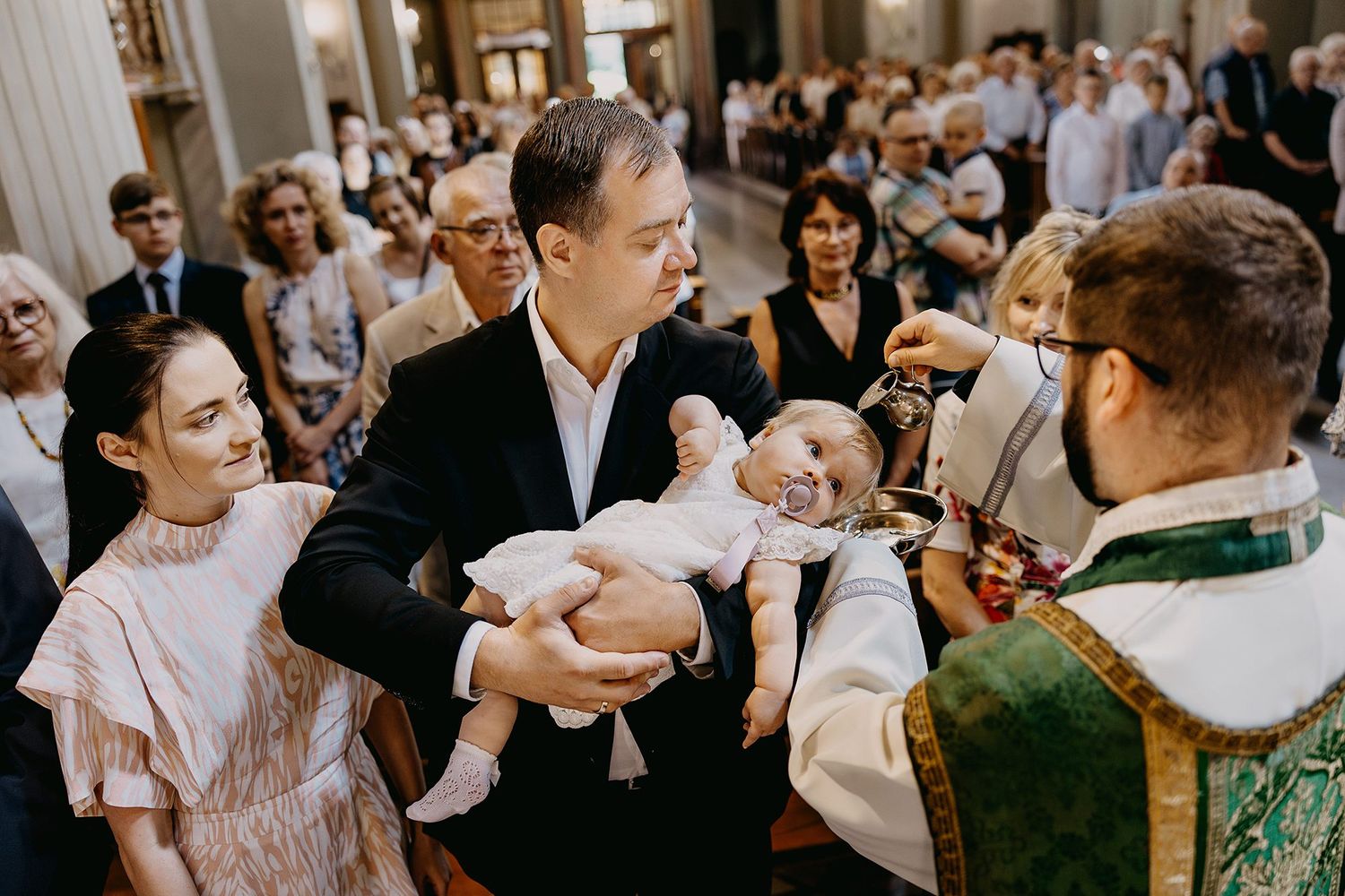 Priest baptizing infant held by father while mother watches during Catholic church ceremony.