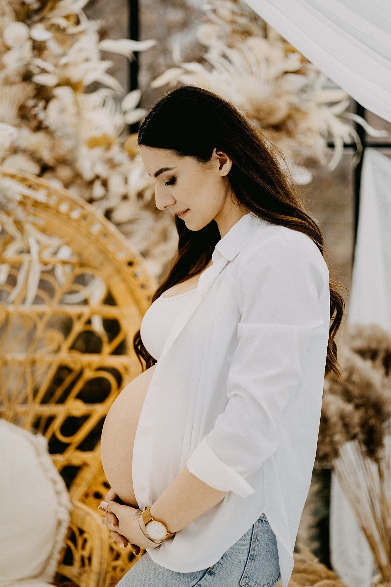 Pregnant woman in white shirt cradling baby bump beside golden boho floral arrangements.