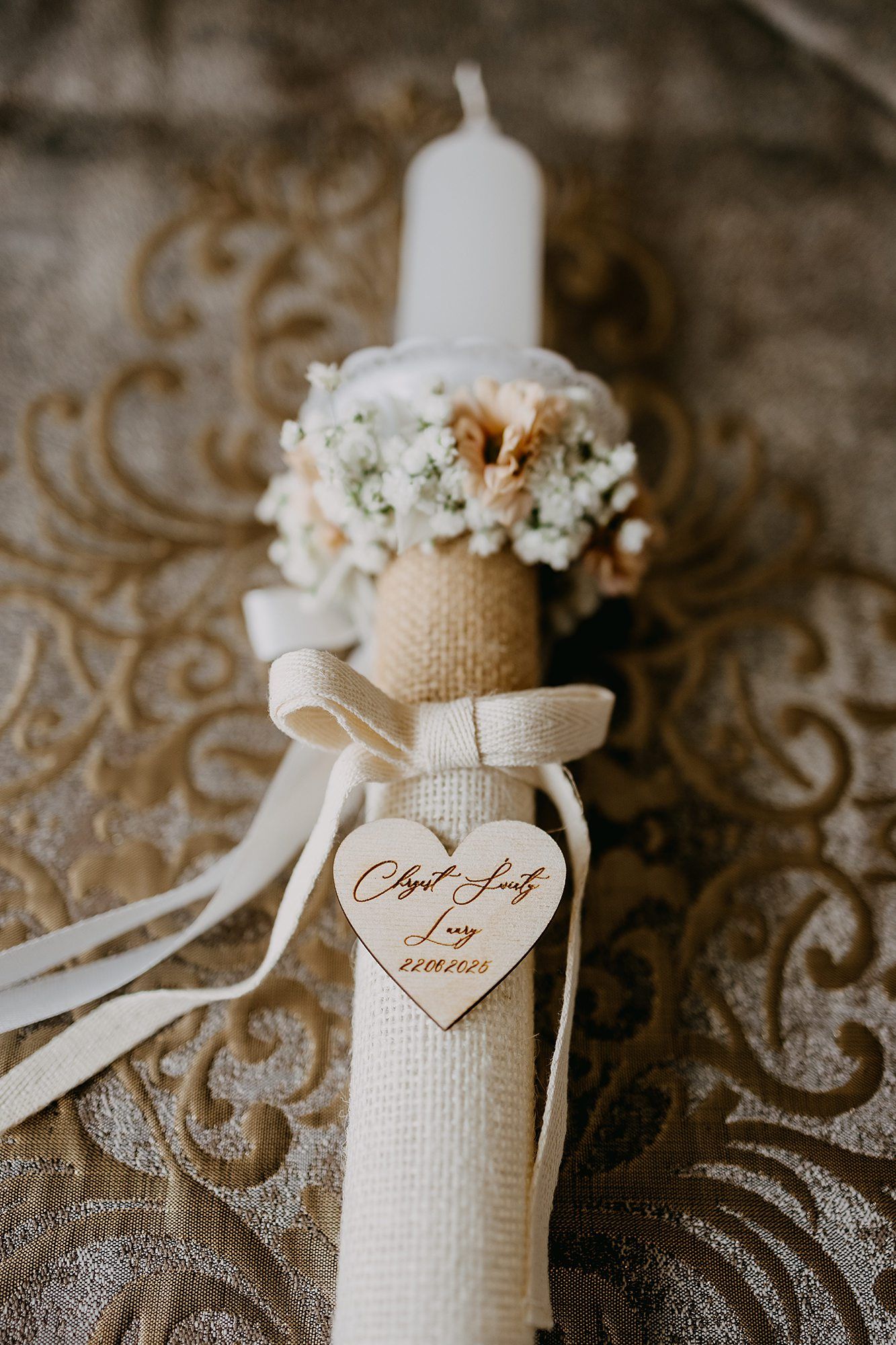 Decorated white baptism candle with burlap, flowers, ribbon bow, and engraved wooden heart christening tag.