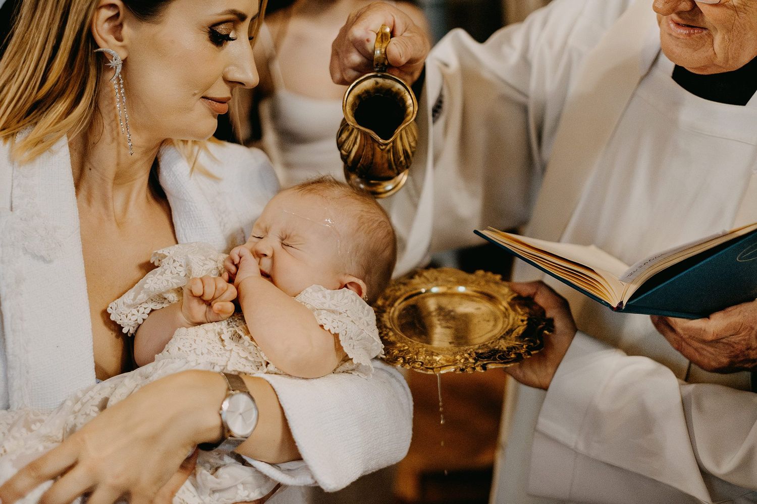 Catholic priest performing infant baptism pouring holy water over newborn held by mother.