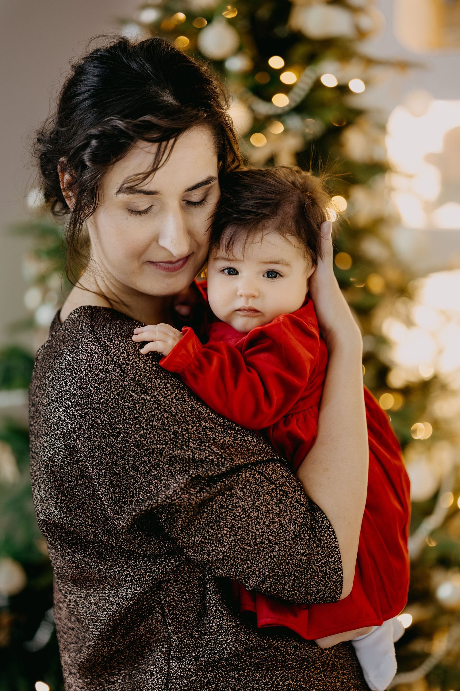 Woman in a sparkly bronze sweater gently holds a baby in a red dress beside a glowing, decorated Christmas tree.