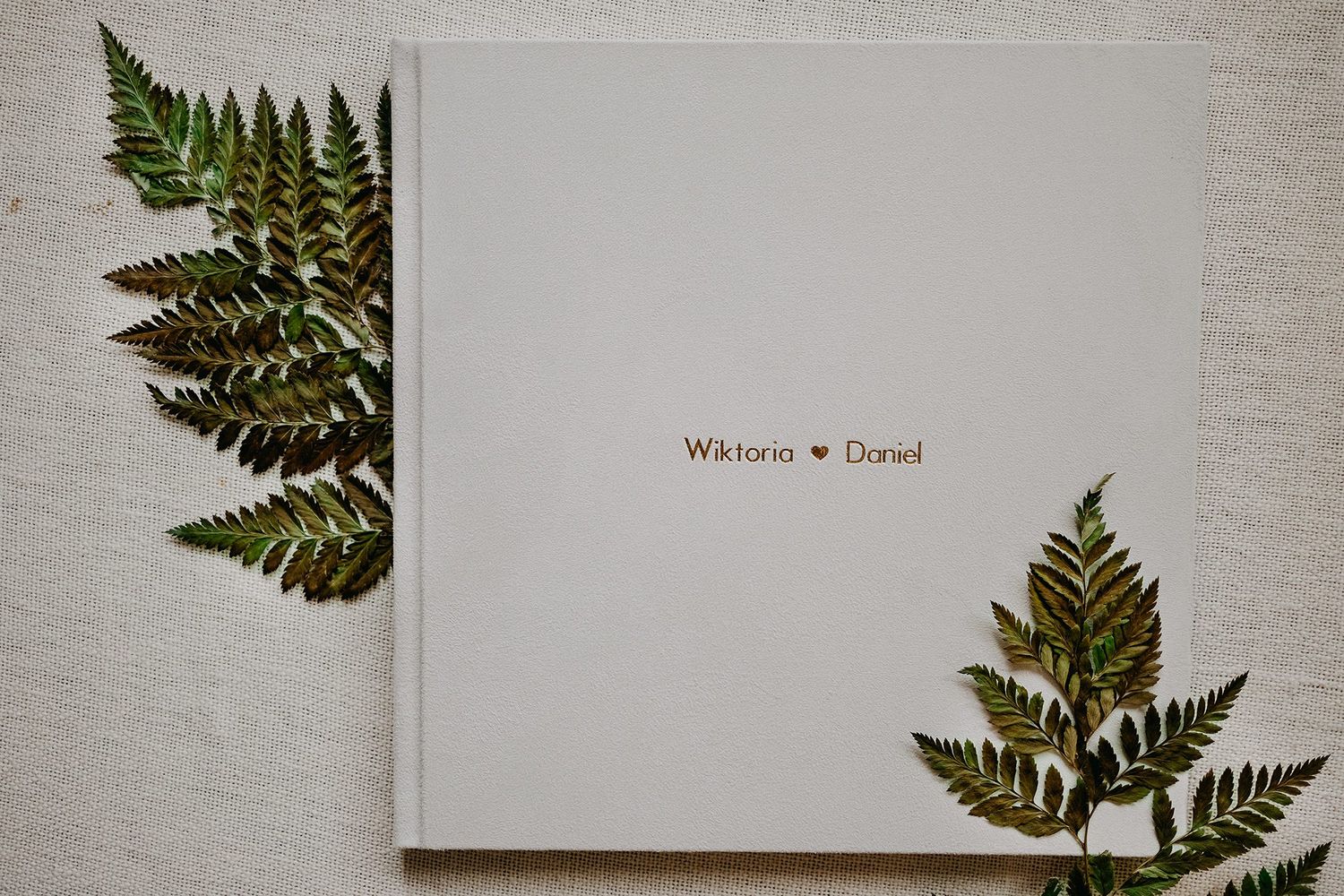 Minimalist white wedding photo album cover with gold text reading Wiktoria and Daniel surrounded by green fern leaves.