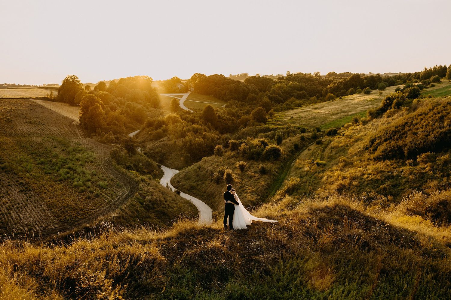 Couple standing on a golden hillside at sunset overlooking a winding river valley surrounded by lush green countryside.