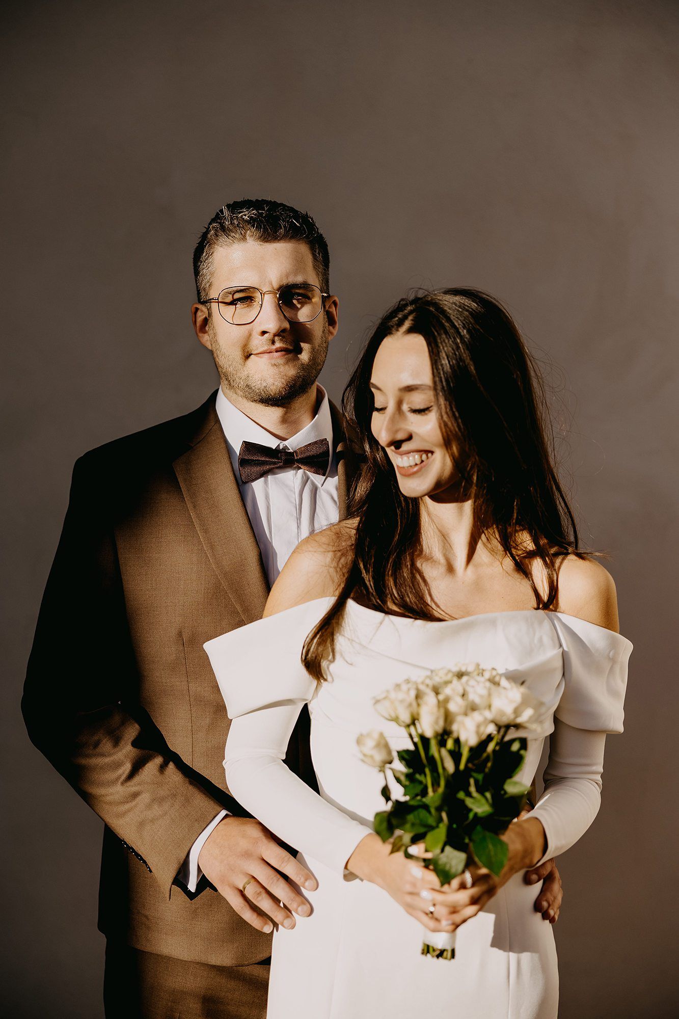 Bride holding white roses bouquet while groom embraces her from behind.