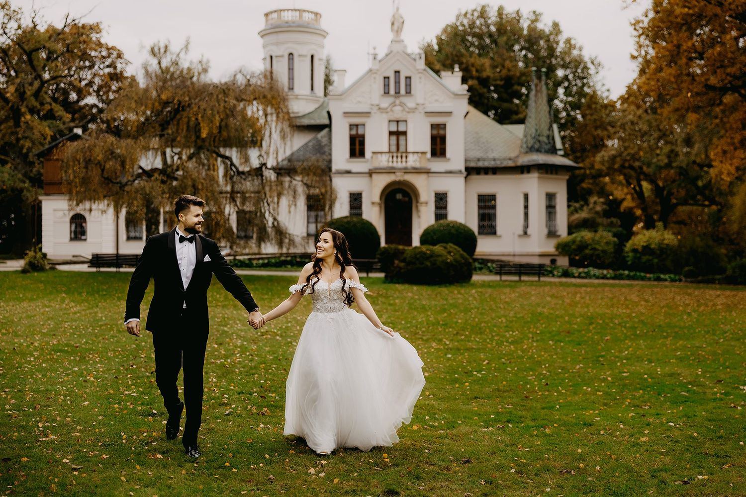 Bride and groom holding hands walking on lawn before elegant manor house in autumn.