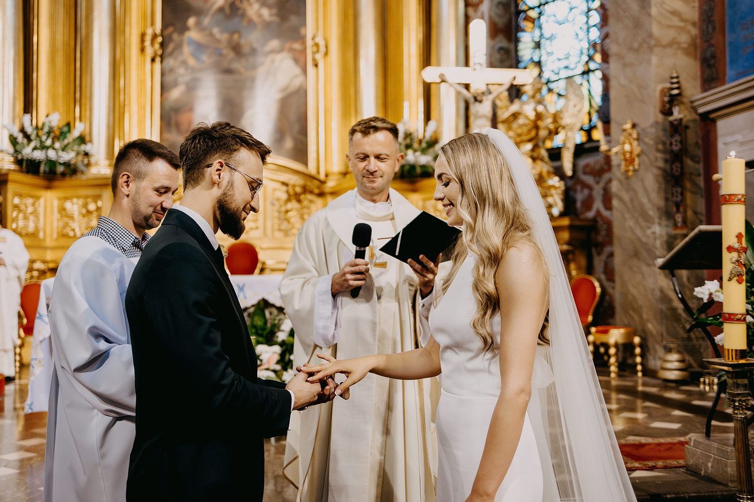 A groom places a ring on the bride's finger during a Catholic church wedding ceremony with a priest.