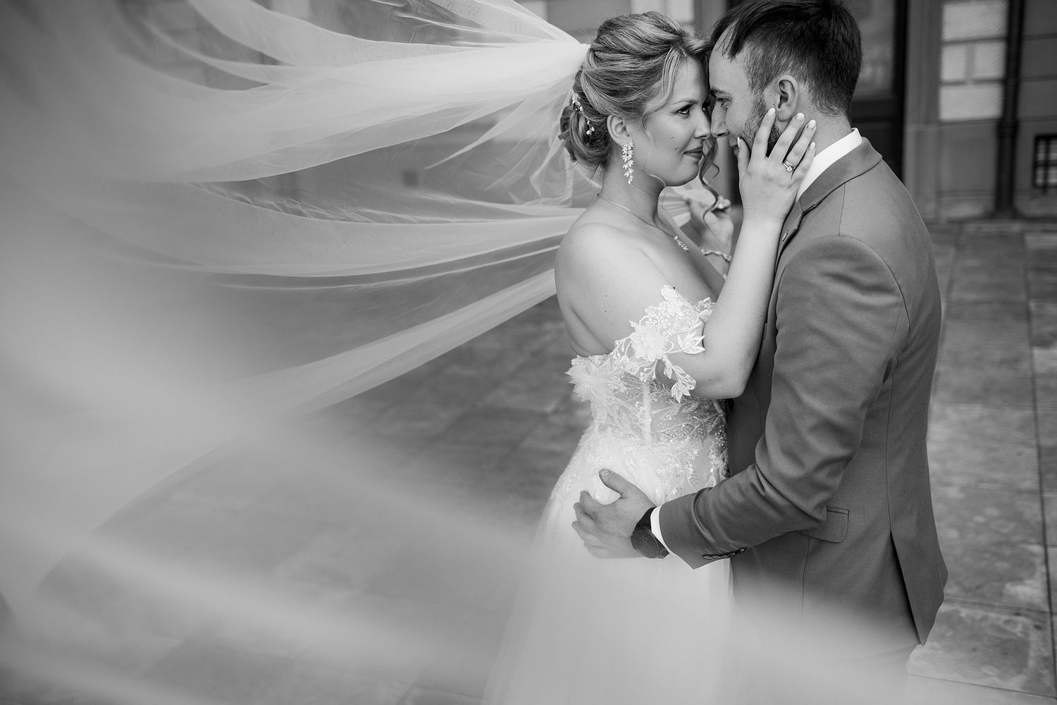 Black and white wedding portrait of couple embracing as bride's veil dramatically billows around them.
