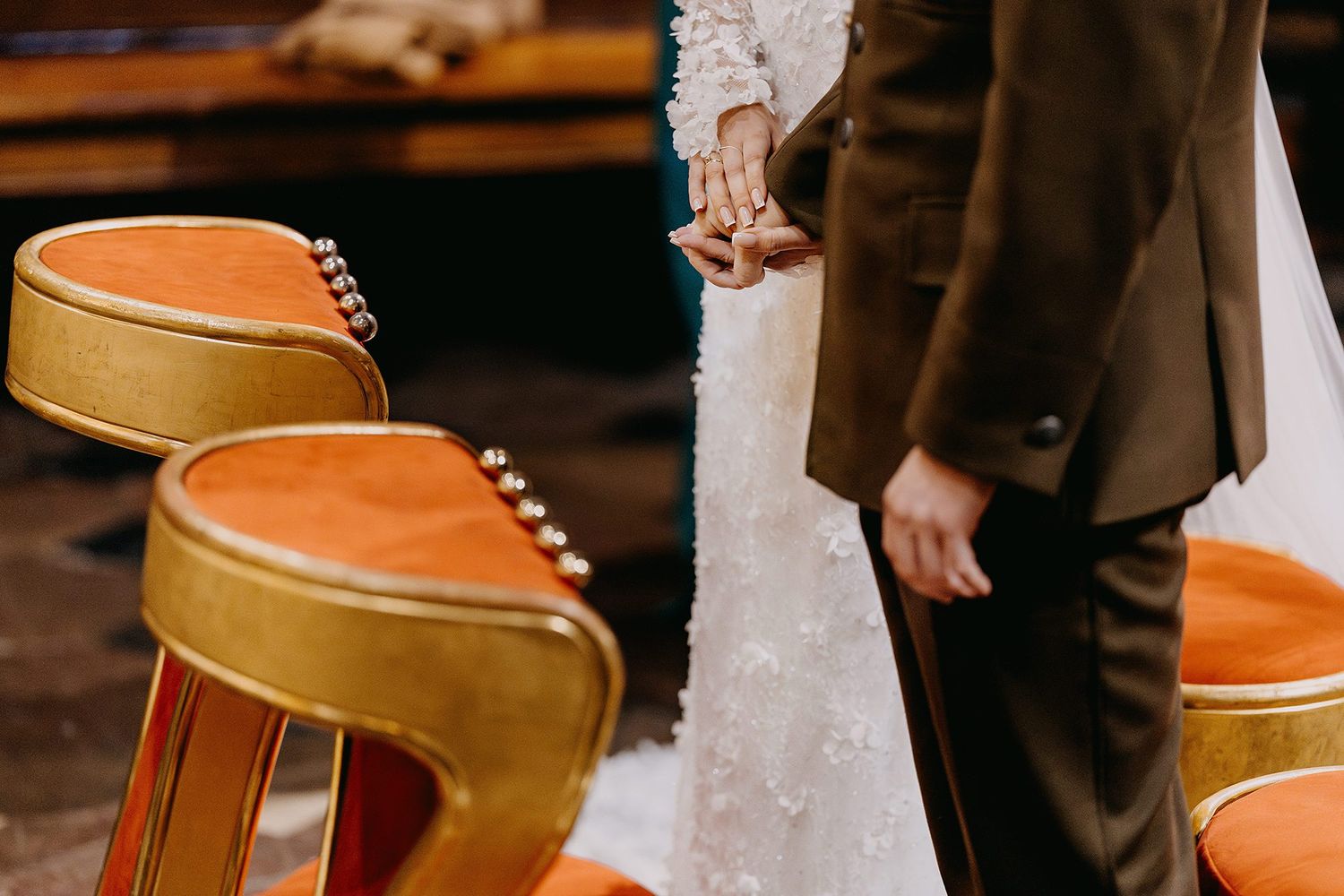 Close-up of bride and groom holding hands near ornate orange velvet church kneelers.