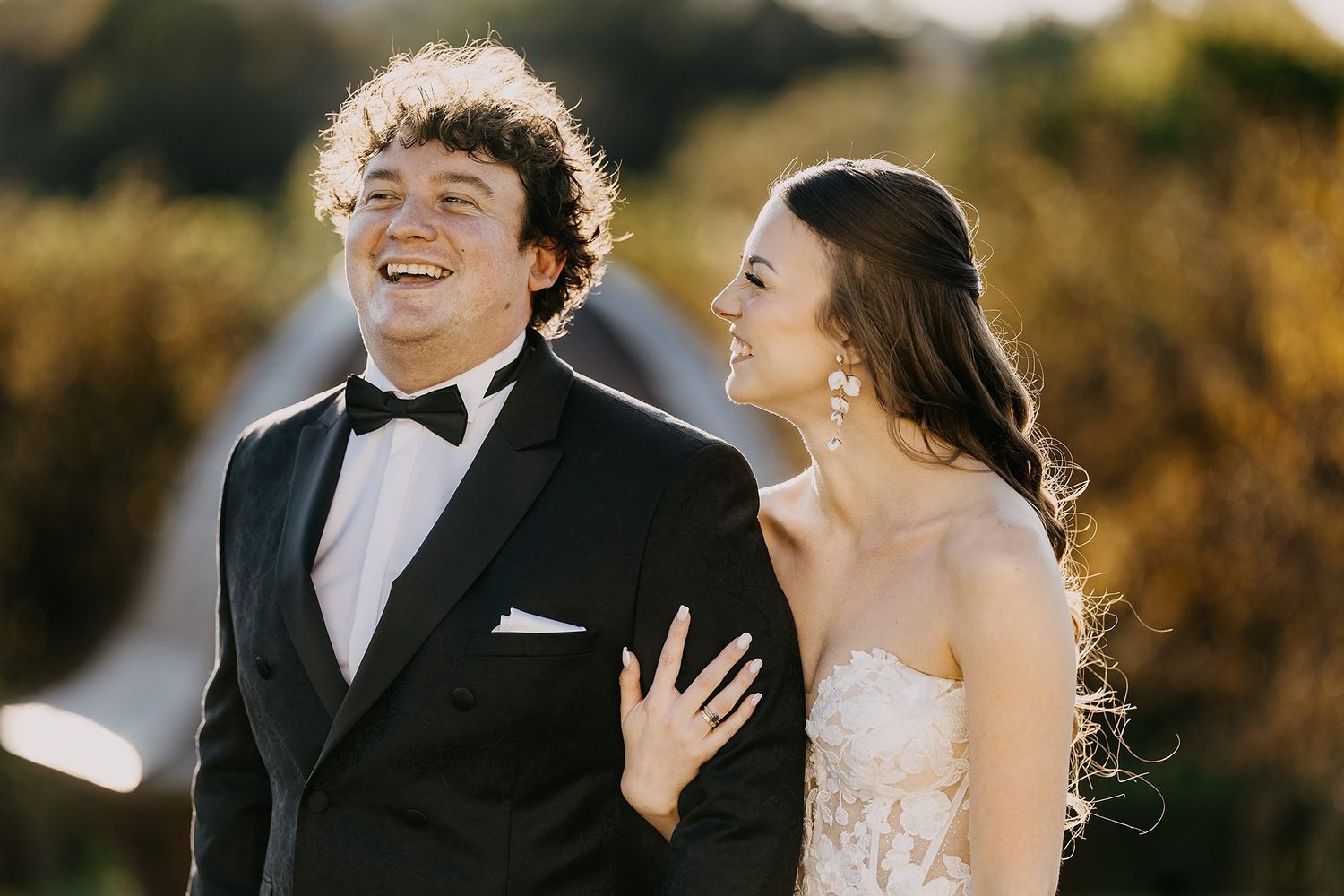 Laughing bride and groom in formal attire share joyful moment outdoors in warm golden light.