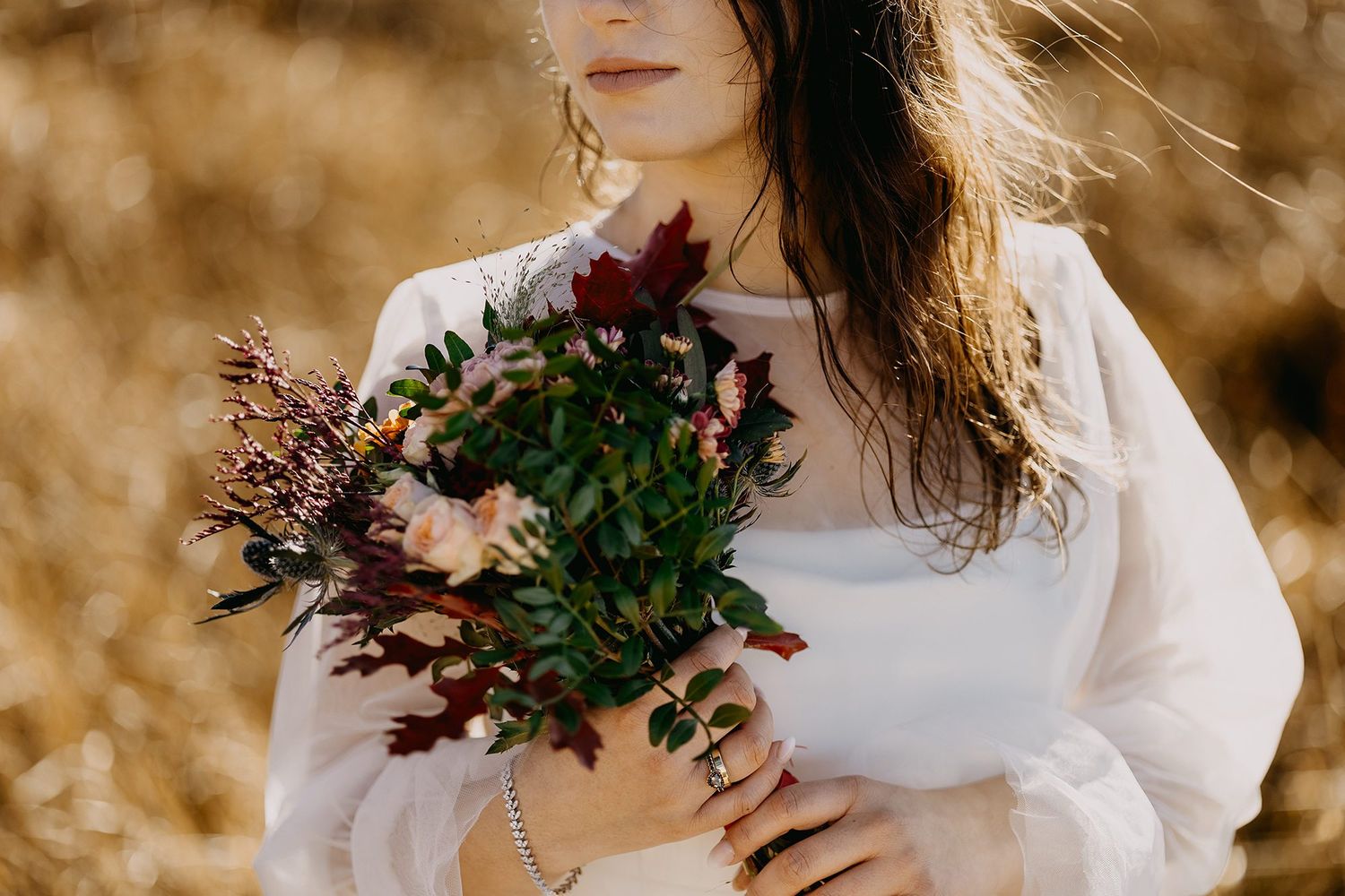 Bride in white dress holding autumn burgundy and green bouquet in golden field setting.