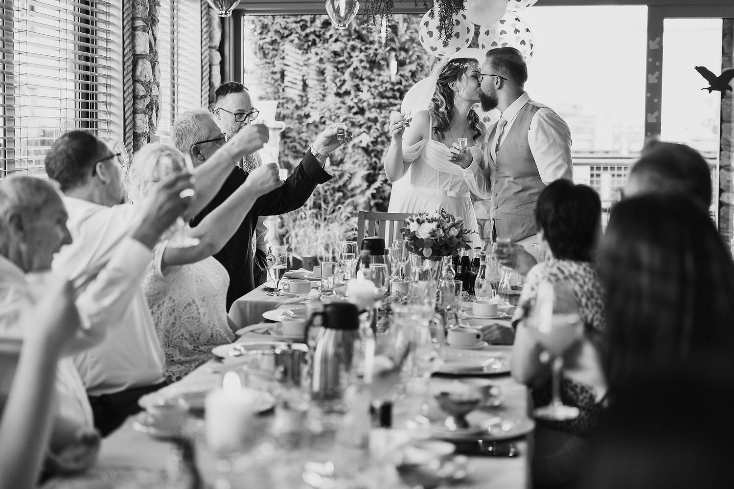 Black and white photo of wedding guests toasting newlyweds kissing at elegant reception dinner.