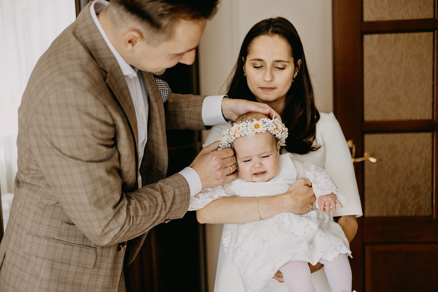 Parents placing floral crown on crying baby dressed in white lace christening gown before baptism ceremony.