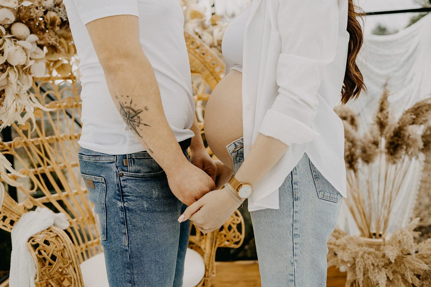 Close-up of couple holding hands, man with tattoo and woman in jeans during maternity shoot.