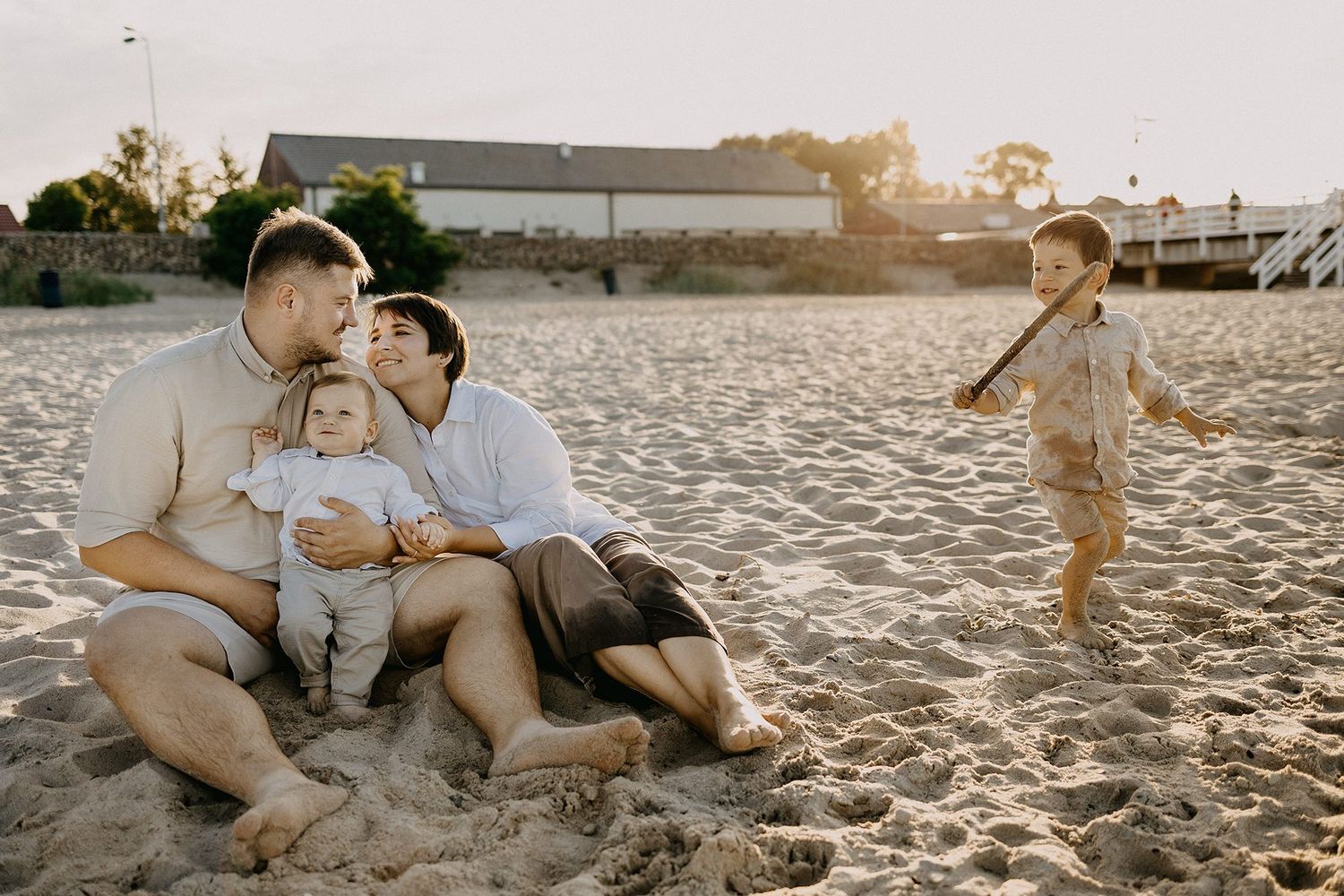 Couple sitting on sandy beach at sunset cuddling baby while young child plays nearby with a stick.