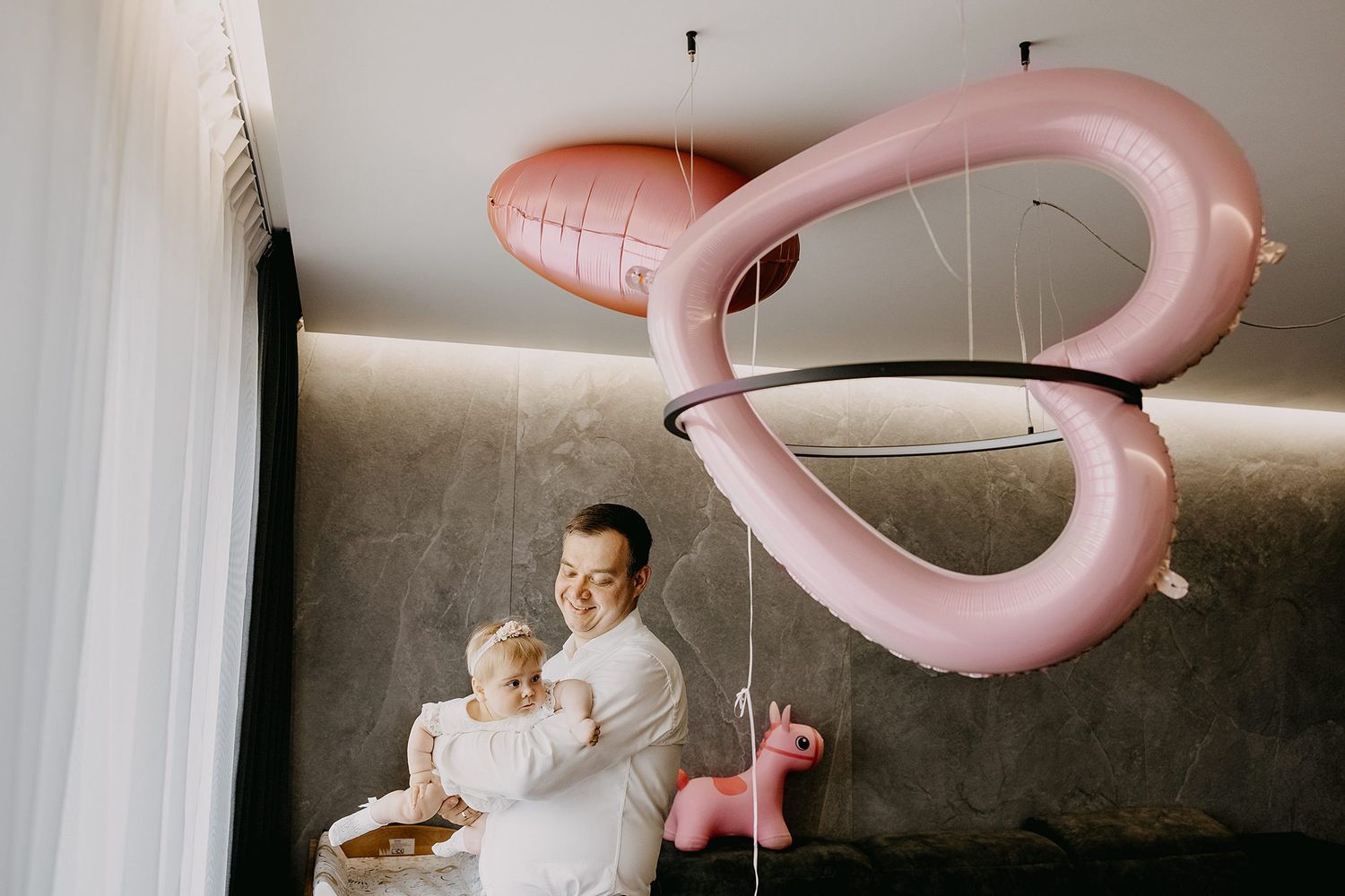 Father holding baby near pink inflatable flamingo balloon decorations in modern interior.