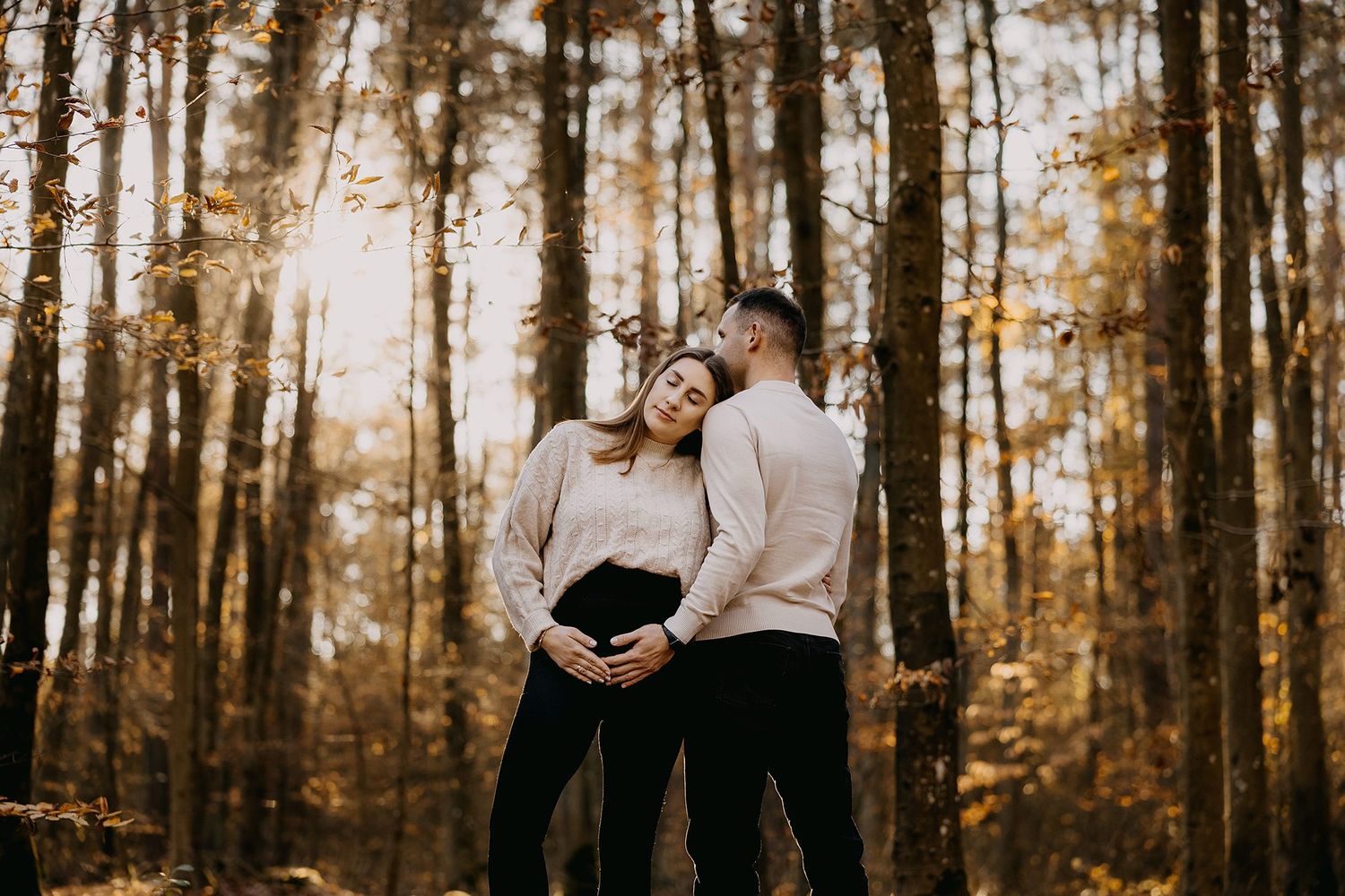Pregnant couple embracing in autumn forest, woman leaning back against partner among golden trees.