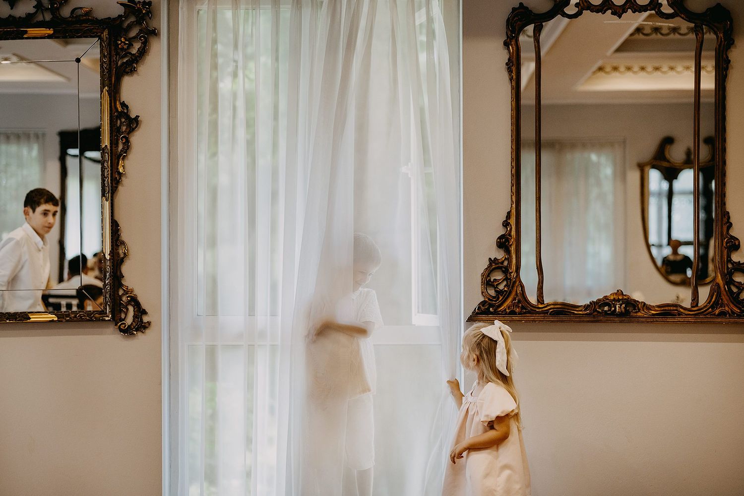 Young girl in white dress standing by sheer curtain with ornate gold mirrors reflecting bride.