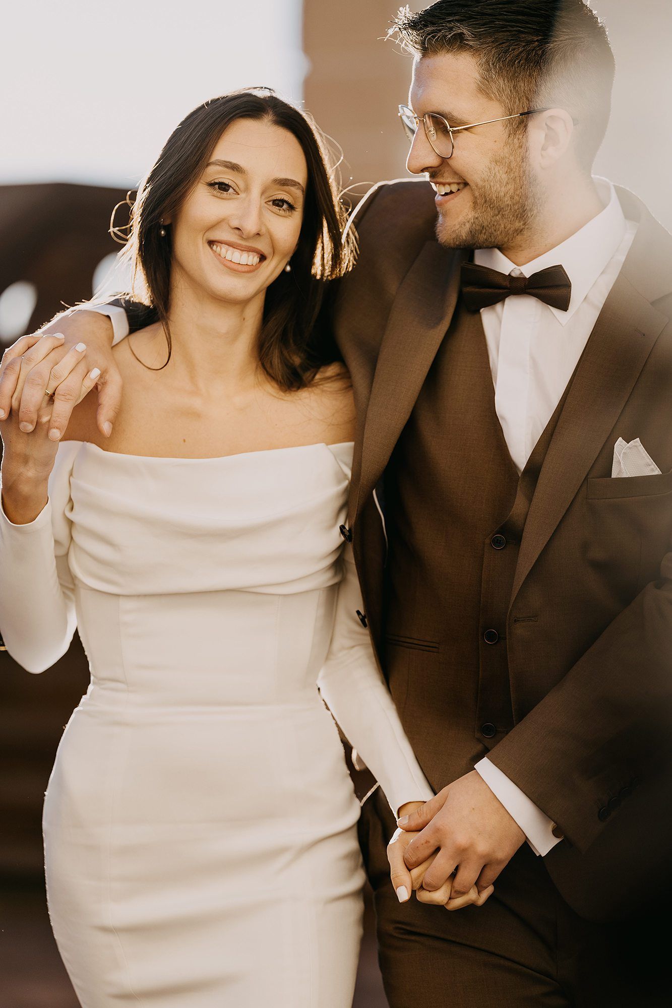 Smiling bride in off-shoulder white dress and groom in brown tuxedo holding hands in warm light.