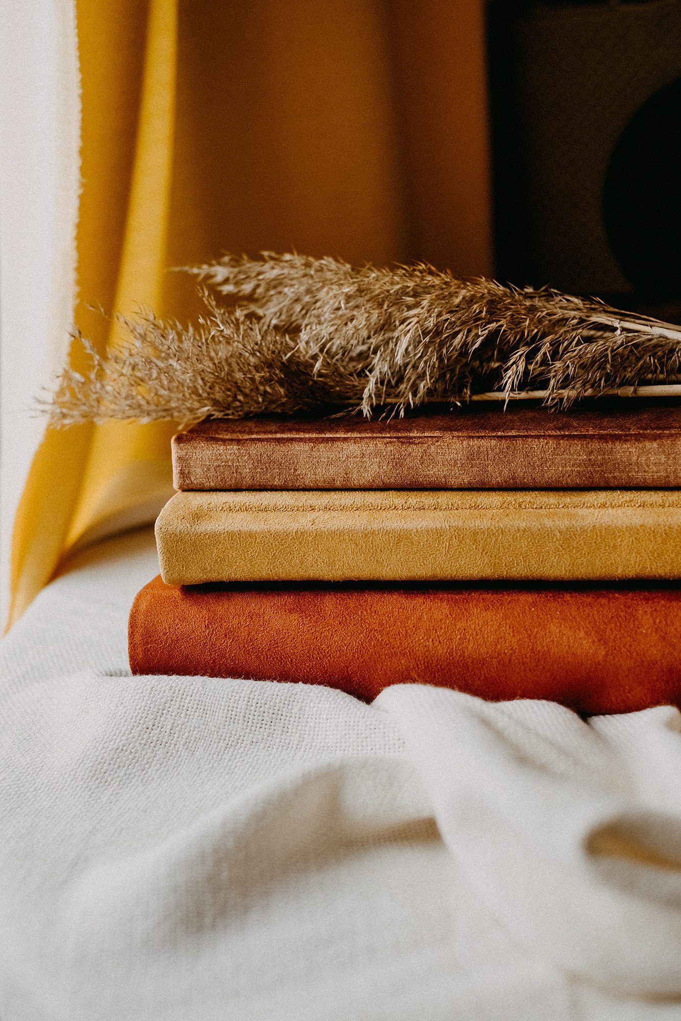 Stack of autumn-toned velvet books with dried pampas grass on white linen near yellow curtain.