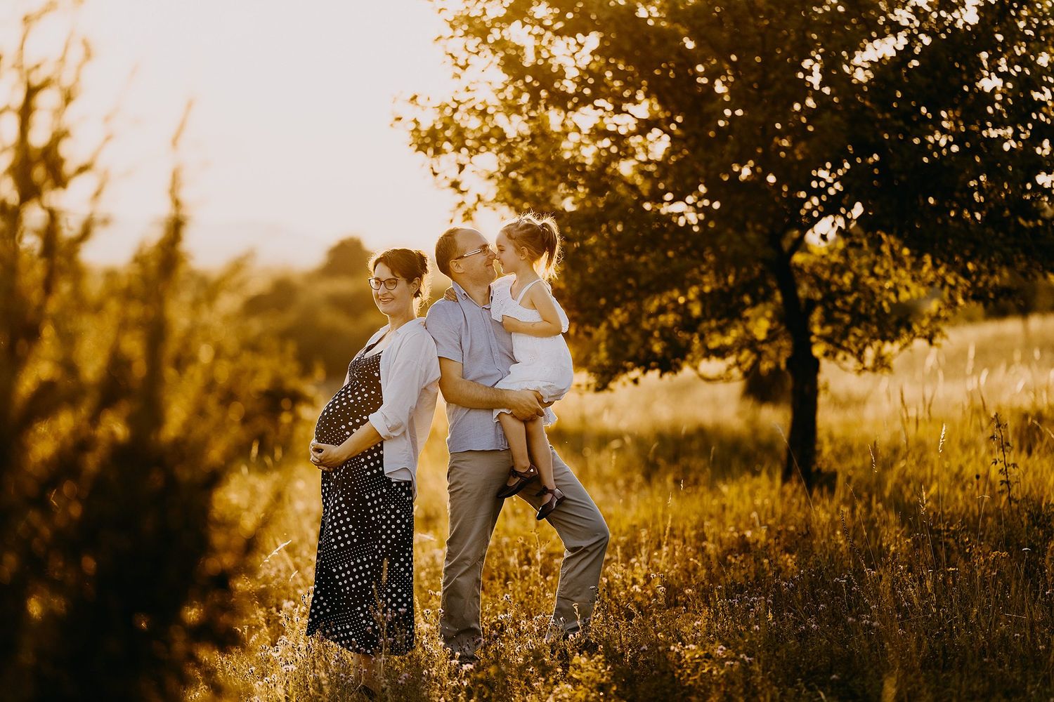 Happy family with pregnant mother, father, and young child sharing a tender moment in golden sunset field.
