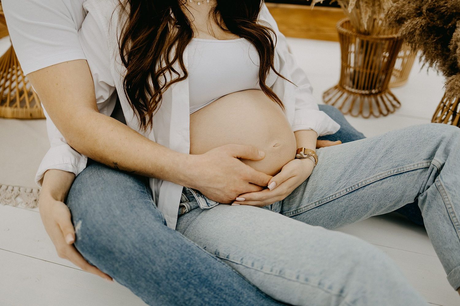 Close-up of couple tenderly holding pregnant belly while sitting together on white floor.