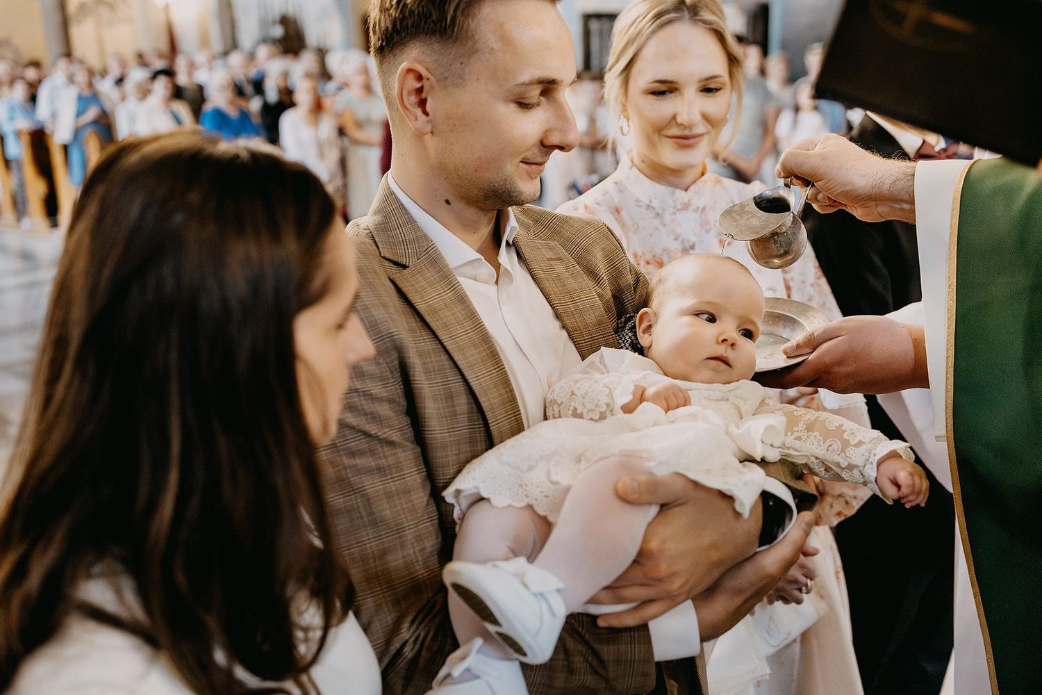 Priest performing Catholic baptism ceremony with parents holding baby in white christening gown at church altar.