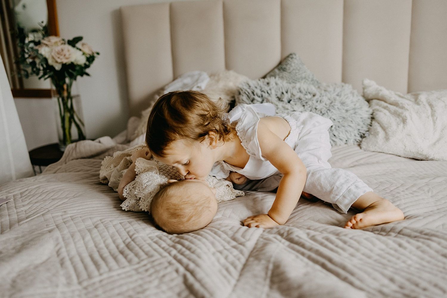 Young toddler gently kisses a newborn baby lying on a soft quilted bed in a cozy bedroom.