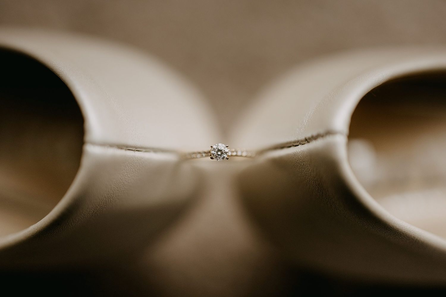 A delicate diamond engagement ring rests between two ivory satin wedding heels in a soft close-up shot.