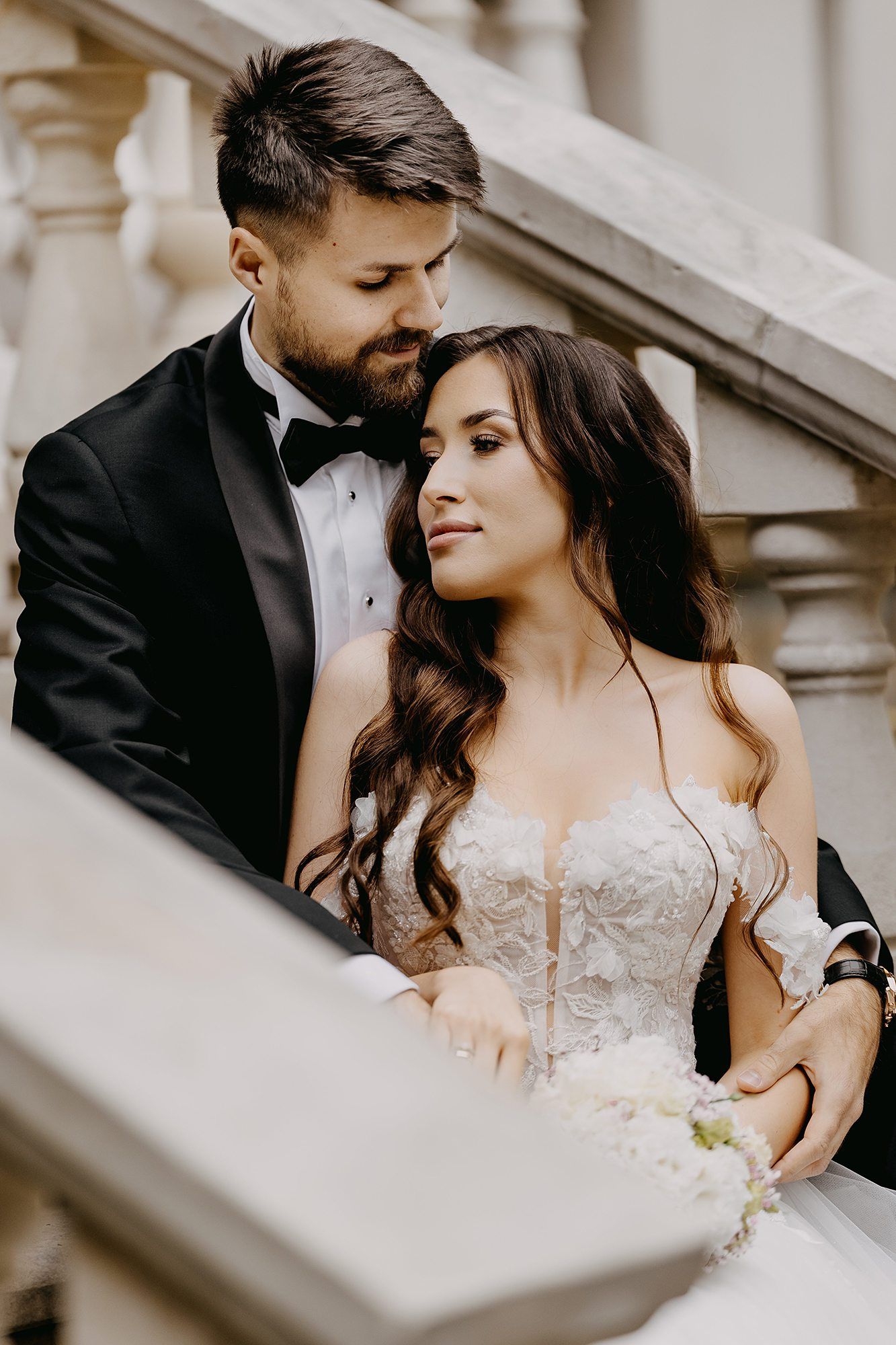 Groom in black tuxedo tenderly embraces bride in strapless lace gown on ornate marble staircase.
