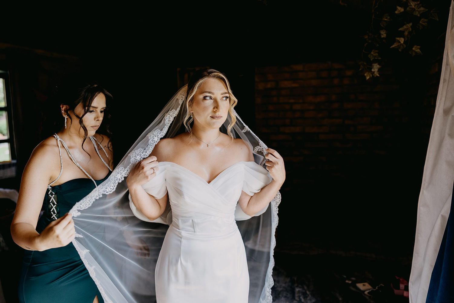 Bridesmaid in teal dress helps bride adjust white lace veil before wedding ceremony.