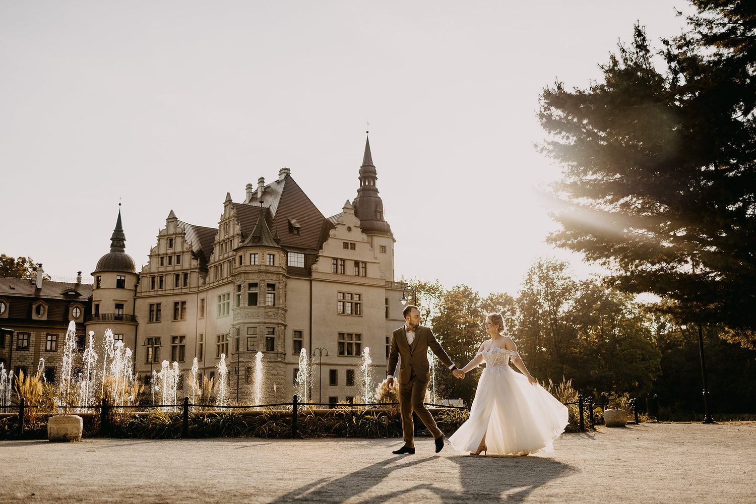 Wedding couple holds hands before a grand castle with fountains glowing in golden sunset light.