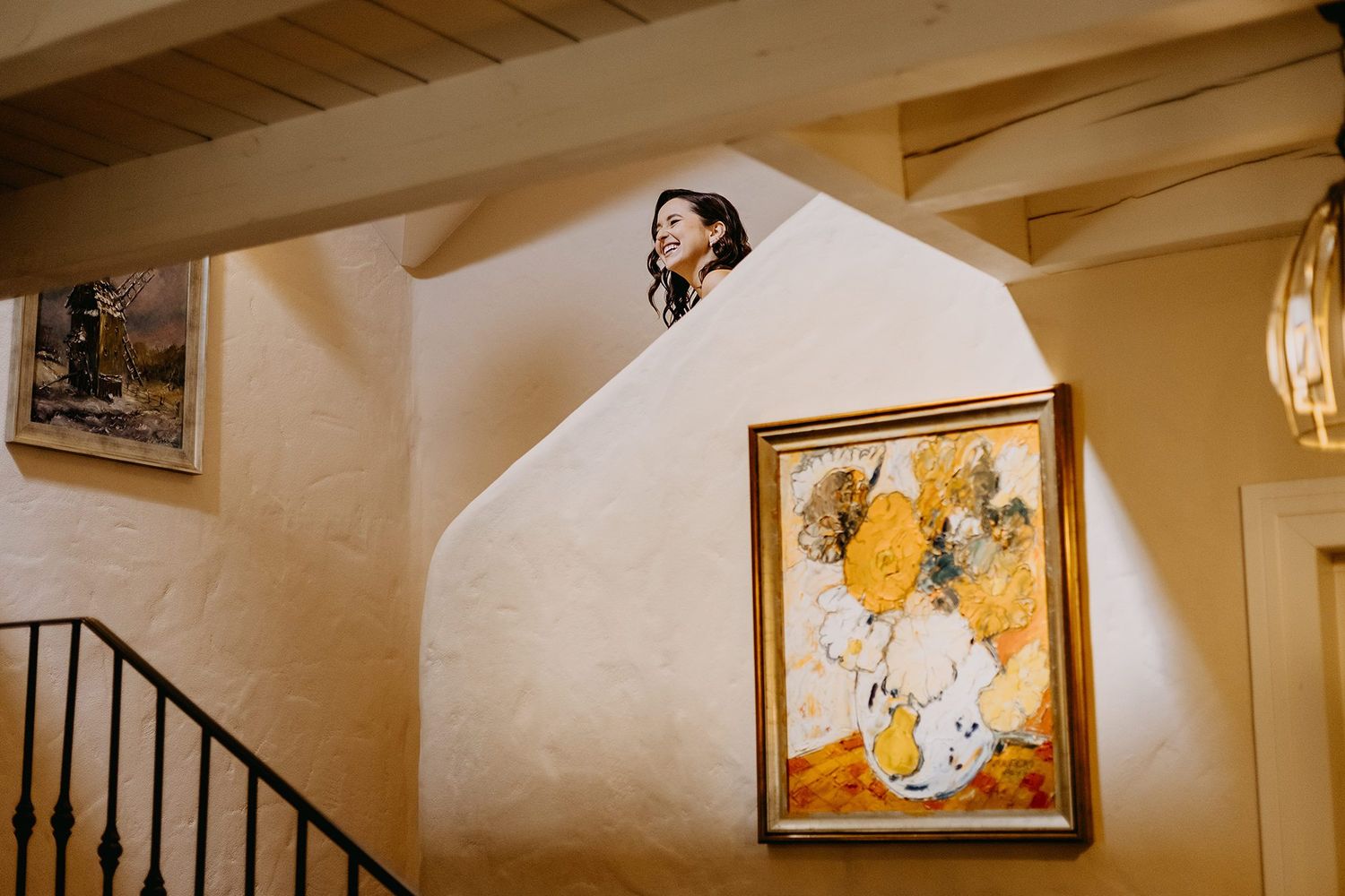 Beautiful bride in white gown peers over a curved staircase wall in a rustic venue.