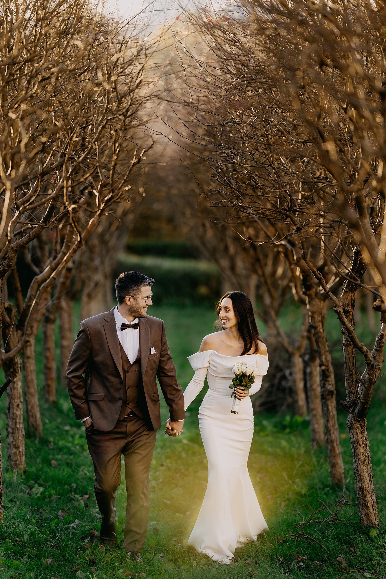 Bride and groom walking hand in hand through a tree-lined garden path.