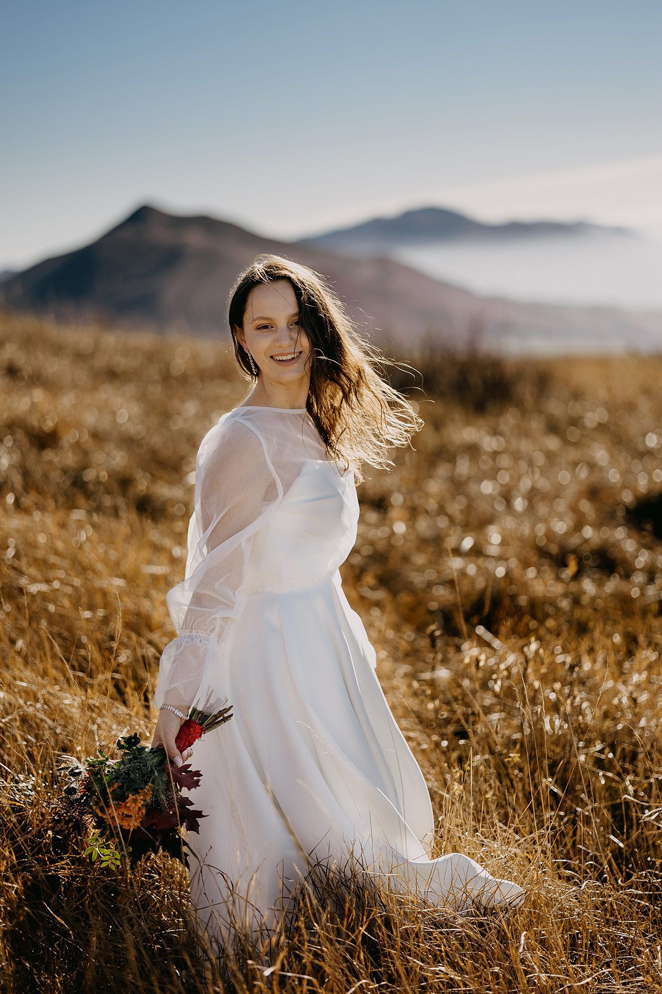 Smiling bride in a white long-sleeve gown standing in a golden meadow with mountain views behind her.
