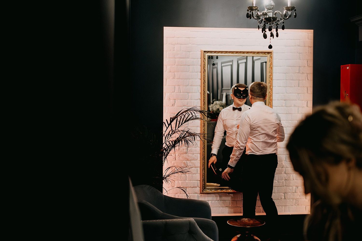 Groom in bow tie adjusting his venetian mask while looking in a decorative mirror at wedding venue.