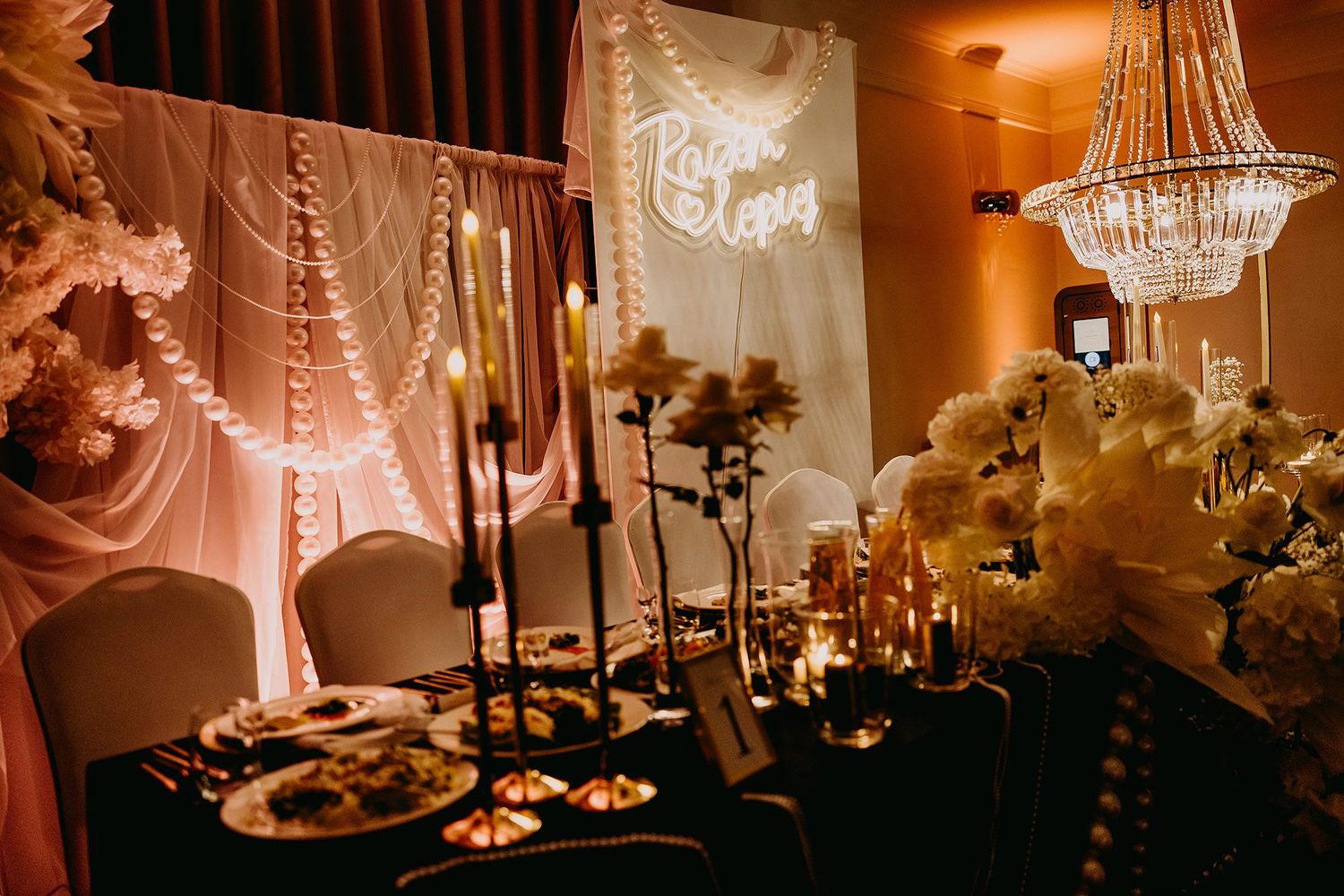 Romantic wedding reception table with candles, white florals, chandelier, and glowing neon name sign.